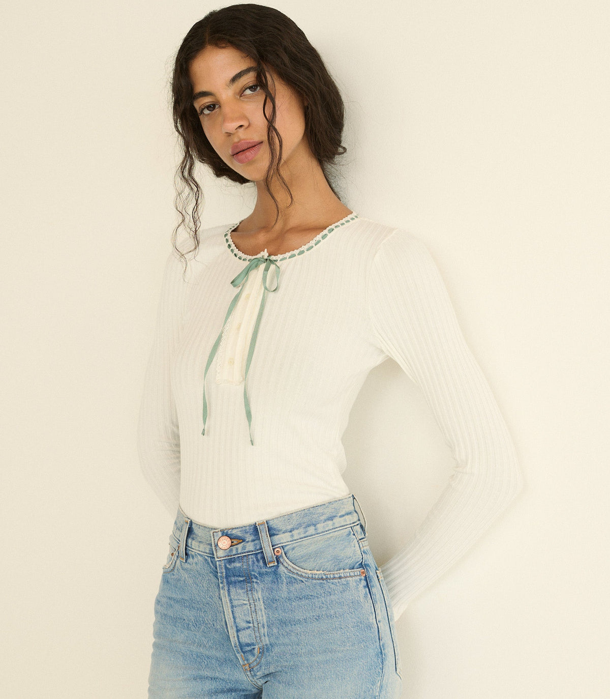 A woman wearing a white ribbed long-sleeve top with a mint blue tie detail at the neckline, paired with light blue jeans, against a plain off-white background.