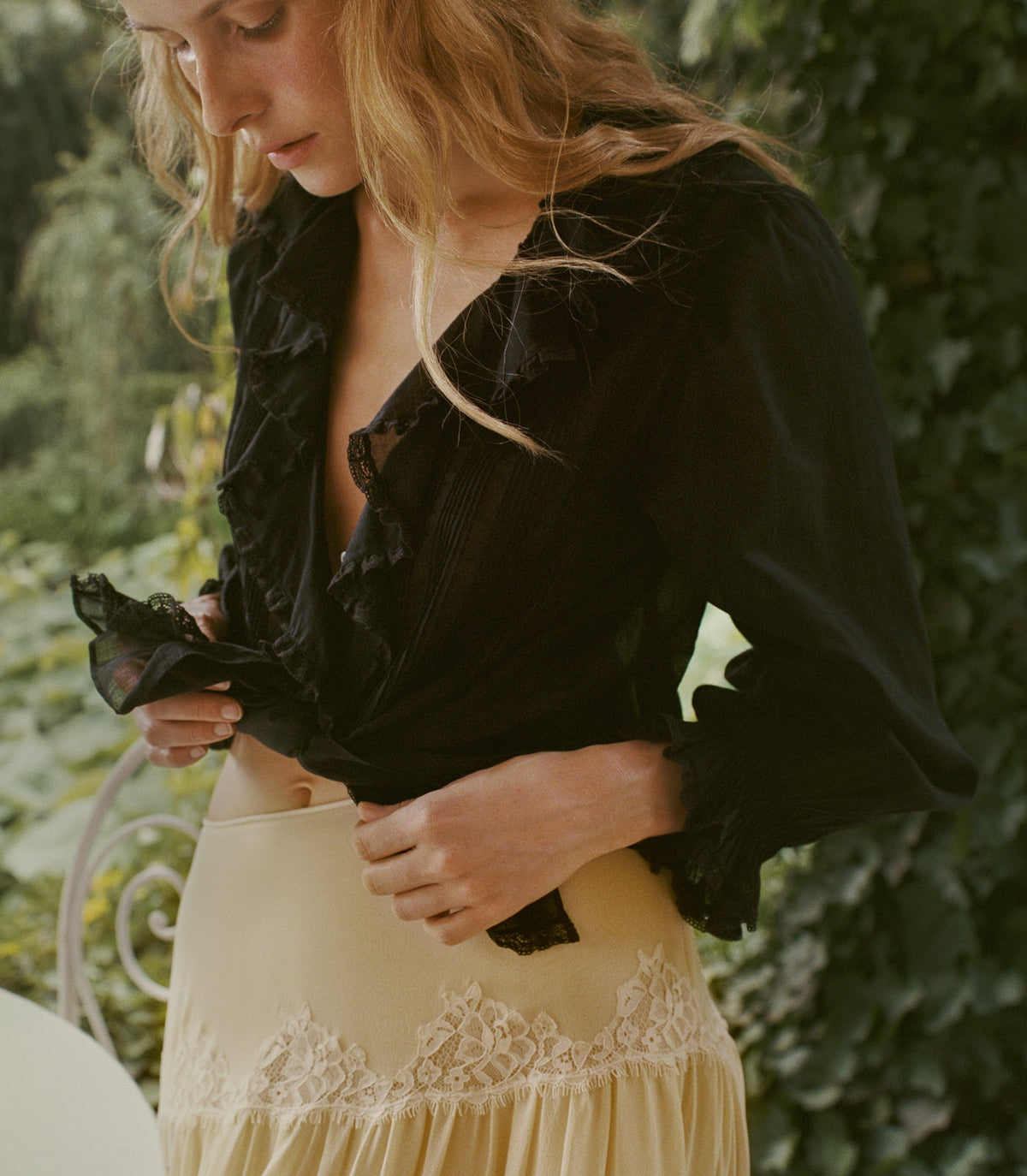 A woman outdoors wearing a black ruffled blouse and a cream lace-trimmed skirt, with green foliage in the background.