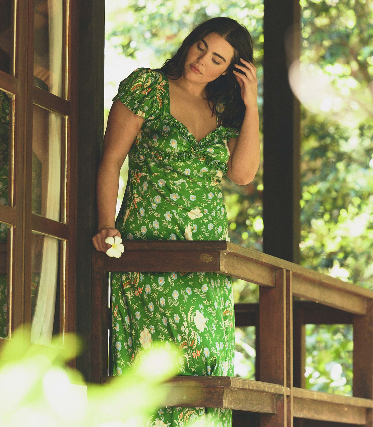 Woman in a green floral dress standing on a wooden deck with greenery in the background